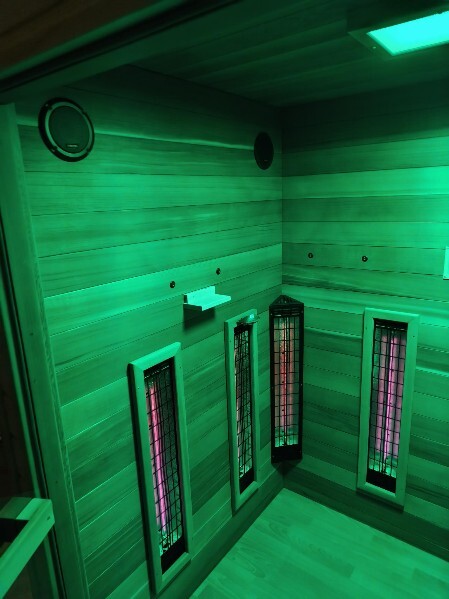 Innenraum der Infrarotsauna „Sophia 160x130“ für Rollstuhlfahrer. Die Wände bestehen aus waagerecht angeordneten Rotzeder-Holzpaneelen. Die Infrarotstrahler sind für Rollstuhlfahrer bodennah montiert und von Schutzgittern abgedeckt. Die Strahlungsintensität ist regelbar und jeder Strahler ist separat zuschaltbar. Die Beleuchtung taucht den Raum in grünes Licht.